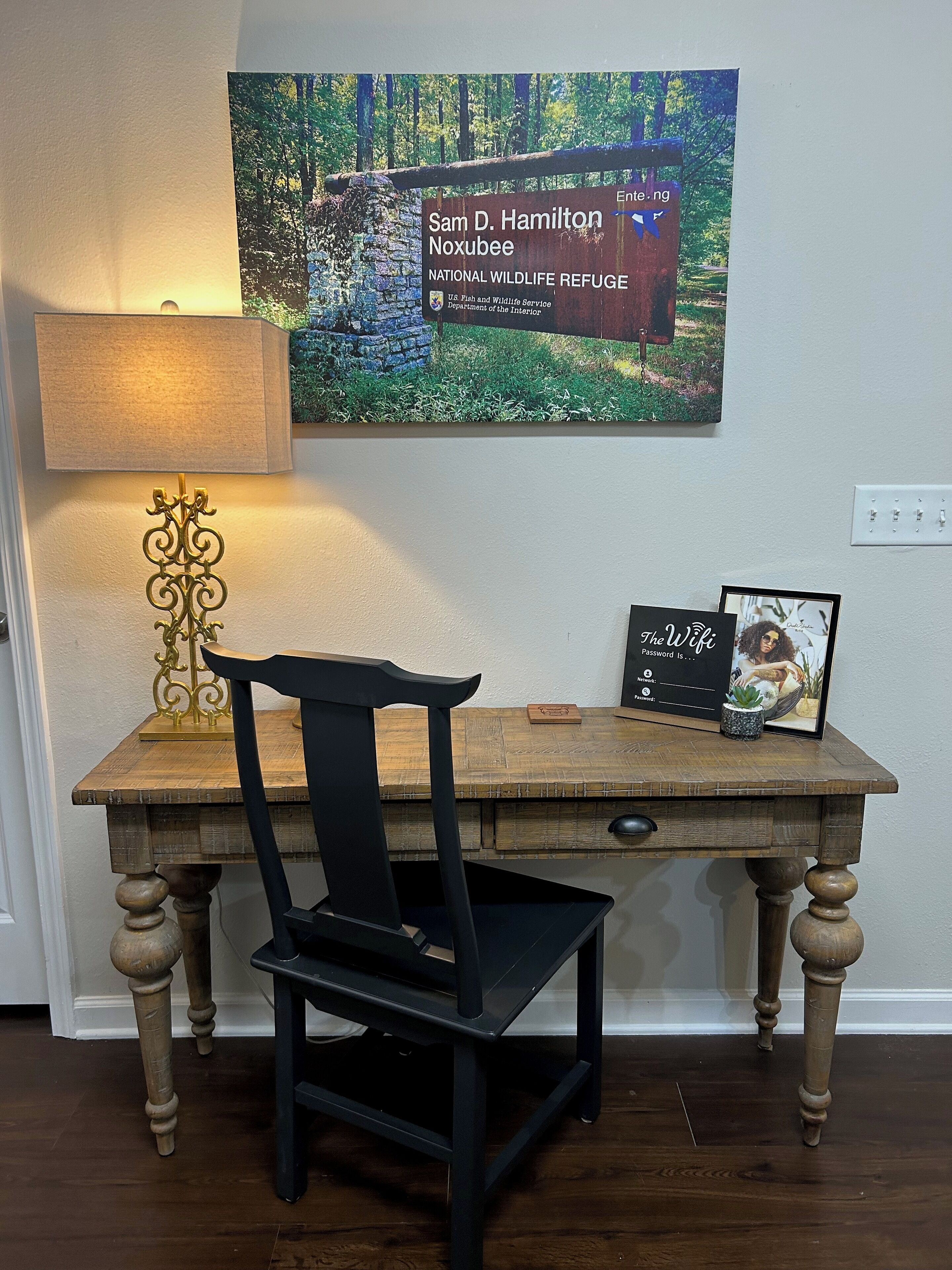 A wooden desk with a black chair, a framed picture, a small potted plant, and a lamp.