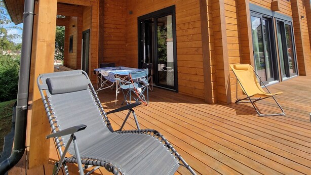 A wooden deck with two lounge chairs, a table, and a glass door leading to a covered patio area. - Eldeweiss Normand (Saint-Marcel)