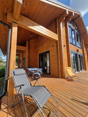 A wooden deck with two lounge chairs, a table, and a glass door leading to a covered patio area. - Eldeweiss Normand (Saint-Marcel)