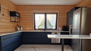 A modern kitchen with wooden walls, a marble floor, a stainless steel refrigerator, and a window with curtains. - Eldeweiss Normand (Saint-Marcel)
