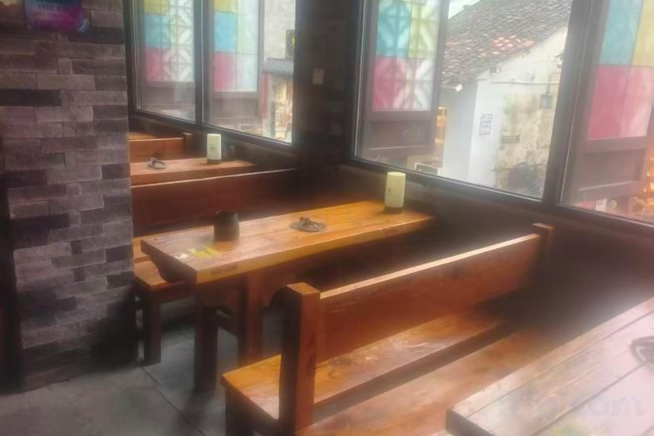 Interior de restaurante com bancos e mesas de madeira, janelas amplas e parede de tijolos.