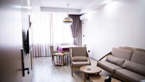 A modern hotel room with a sofa, armchair, coffee table, and a dining area with a table and chairs.