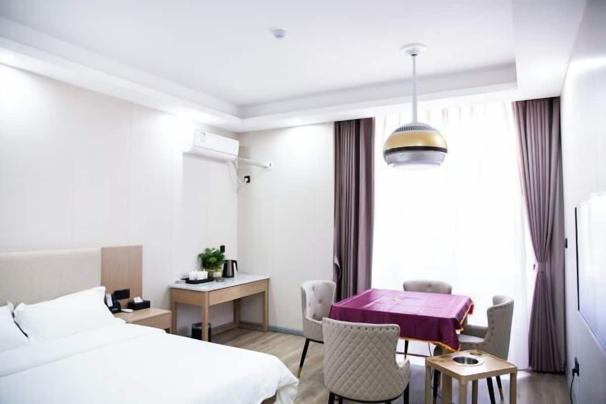 A hotel room with a bed, a desk, a table with chairs, a hanging light fixture, and a window with curtains.