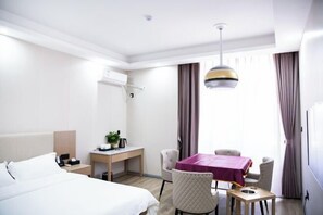 Habitación de hotel con cama, escritorio, mesa con sillas, lámpara colgante y ventana con cortinas.