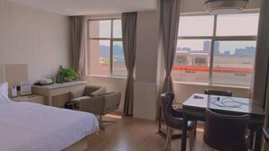 A hotel room with a bed, a desk with a chair, a dining table with chairs, and large windows with curtains.