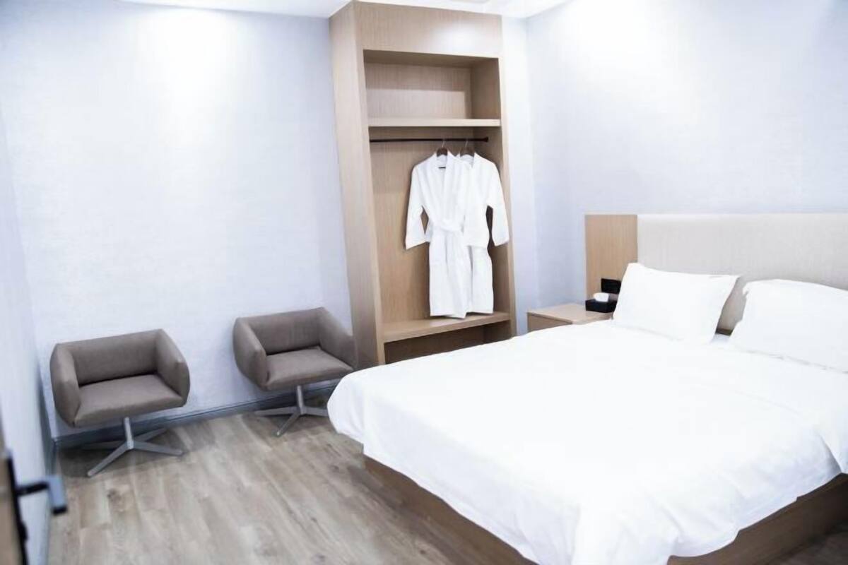 A hotel room with a bed, a seating area with two chairs, a wooden wardrobe, and a bedside table.