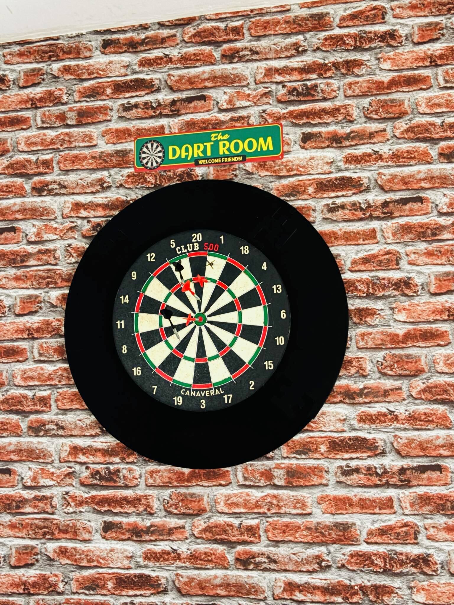 Eine Dartscheibe, an einer Ziegelwand befestigt, mit einem Schild darüber, auf dem 'The DART ROOM' steht.
