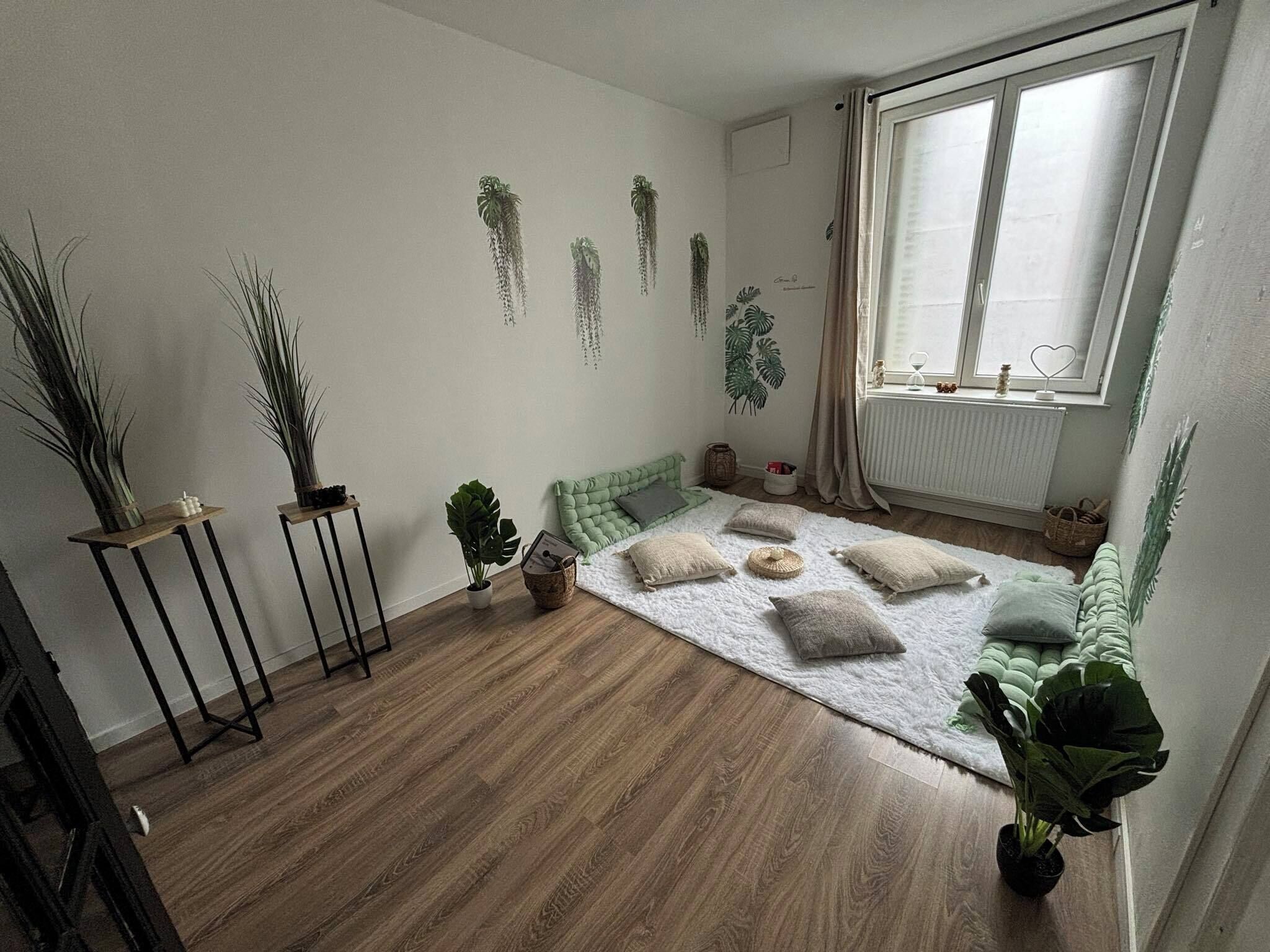 Une pièce avec un plancher en bois, un tapis blanc et un canapé vert. On y trouve plusieurs plantes en pot et des œuvres d’art accrochées au mur.