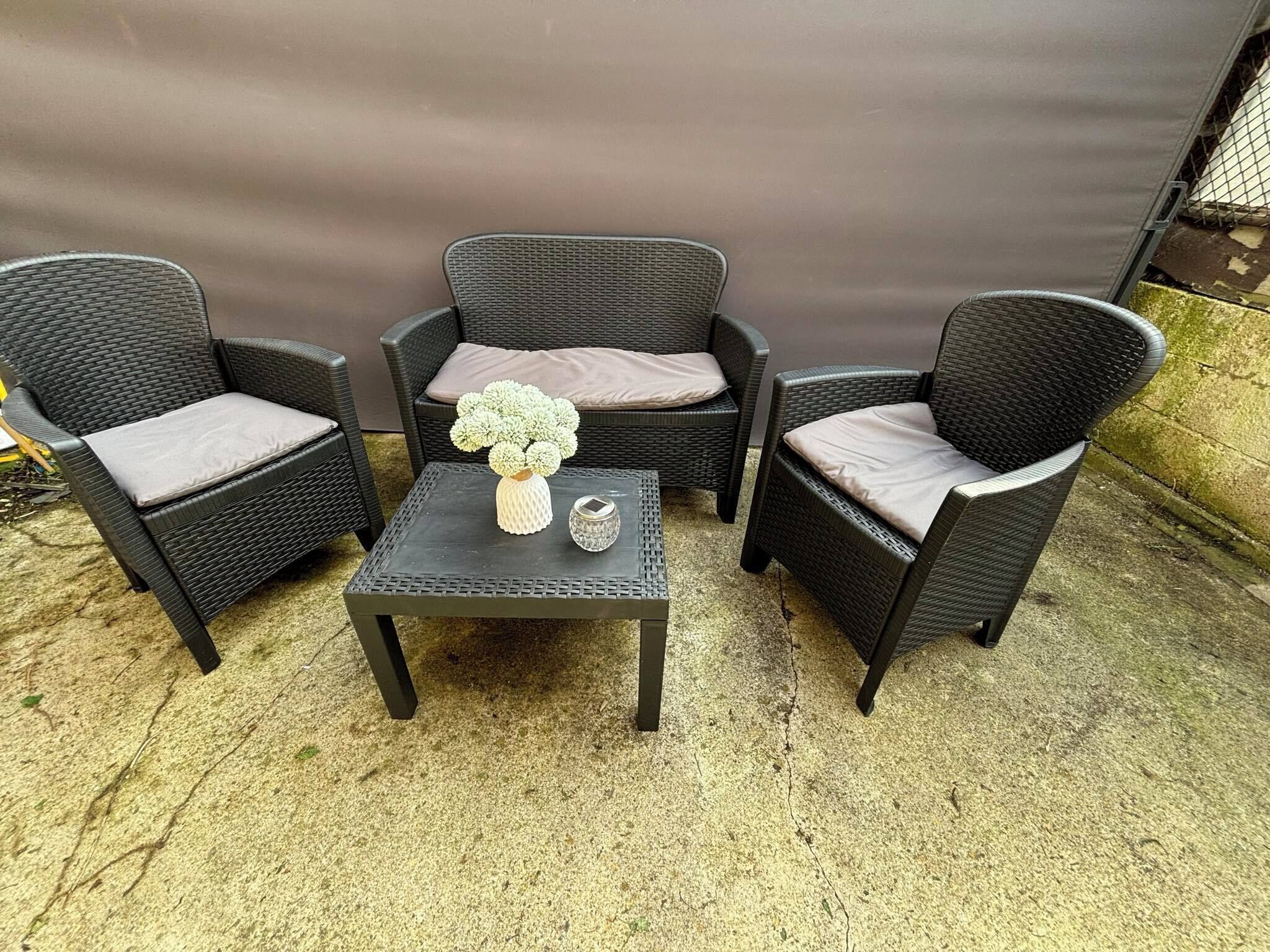 A set of three wicker chairs with cushions, a small table with a vase and a decorative item, and a concrete floor.