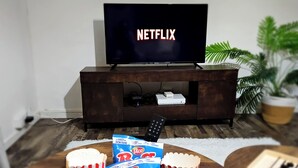 Una sala de estar con un televisor mostrando 'NETFLIX', una mesa de centro de madera con palomitas de maíz y una bolsa de aperitivos, y un control remoto.