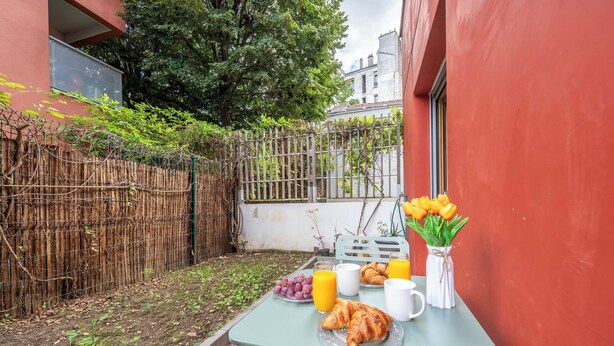 A small outdoor dining area with a table set for breakfast, including croissants, grapes, orange juice, and a vase of tulips.