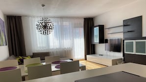 Ein modernes Wohnzimmer mit Essbereich, einem großen Fenster mit transparenten Vorhängen, einer dekorativen Pendelleuchte, einem an der Wand montierten Flachbildfernseher und einer integrierten Unterhaltungsanlage.