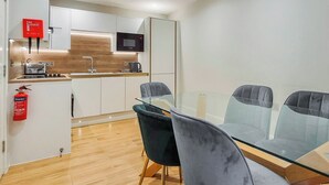 Eine moderne Küche mit weißen Schränken, einer Mikrowelle und einem Toaster. Ein Glastisch mit vier Stühlen steht bereit. Ein Feuerlöscher ist an der Wand angebracht.