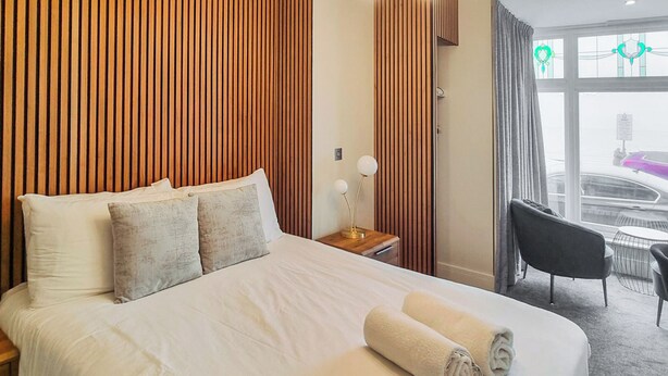 Ein Hotelzimmer mit einem großen Bett, einer Holzwand, einem Nachttisch mit Lampe, einem Sessel und Blick auf die Straße durch das Fenster.