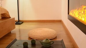 A cozy room with a wooden floor, a round woven rug, a small table with a teapot and cups, and a wall-mounted fireplace.