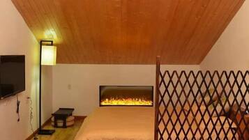 Un dormitorio con techo inclinado, un televisor fijado a la pared, una mesita de noche, una cama con cabecero y una chimenea.