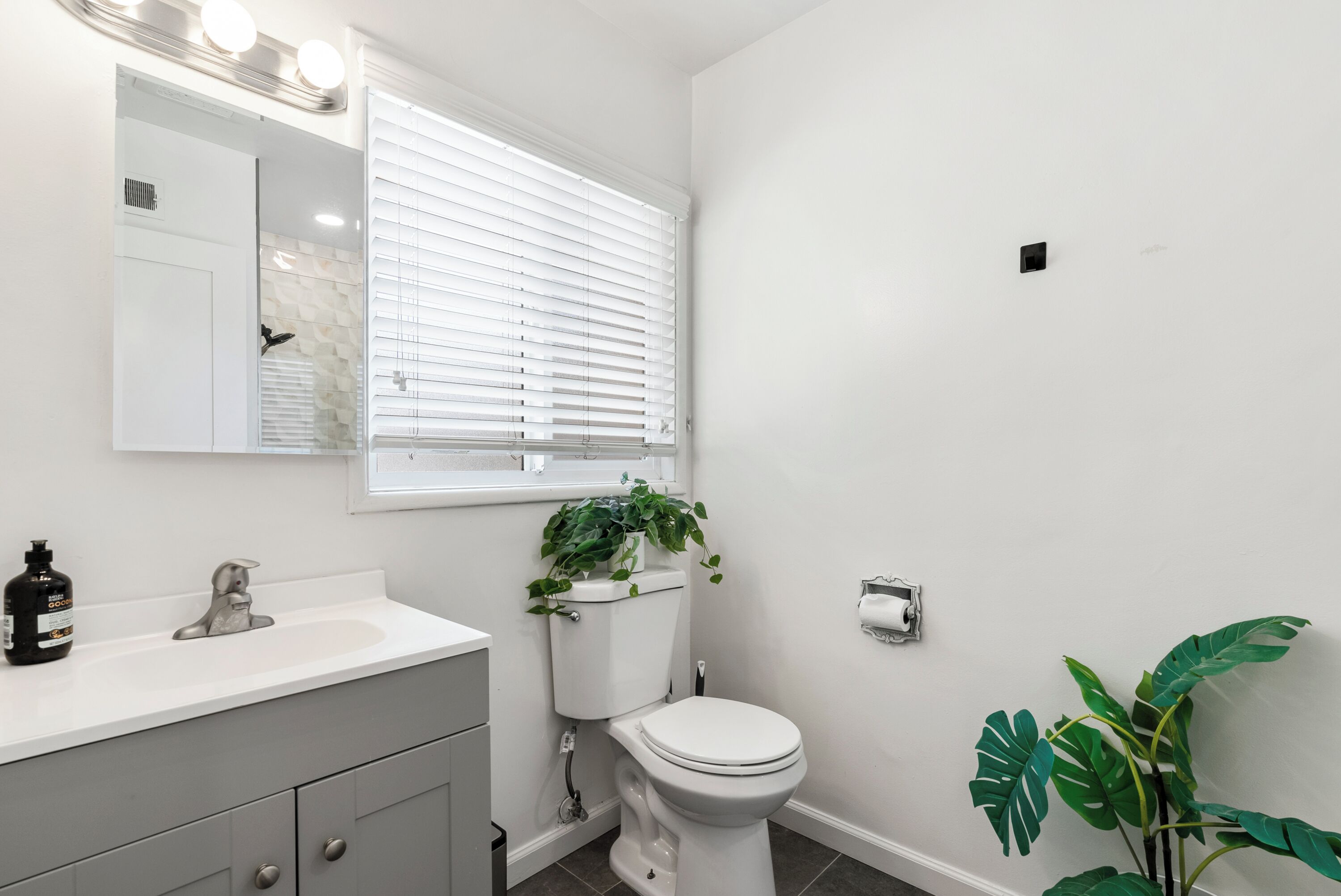 Un baño moderno con un lavamanos, tocador y retrete blancos. Hay una ventana con persianas, un espejo y una planta en maceta.