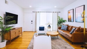 Un salon moderne avec un téléviseur à écran plat, un canapé en cuir marron, un fauteuil blanc, une table basse ronde en bois et deux plantes en pot.