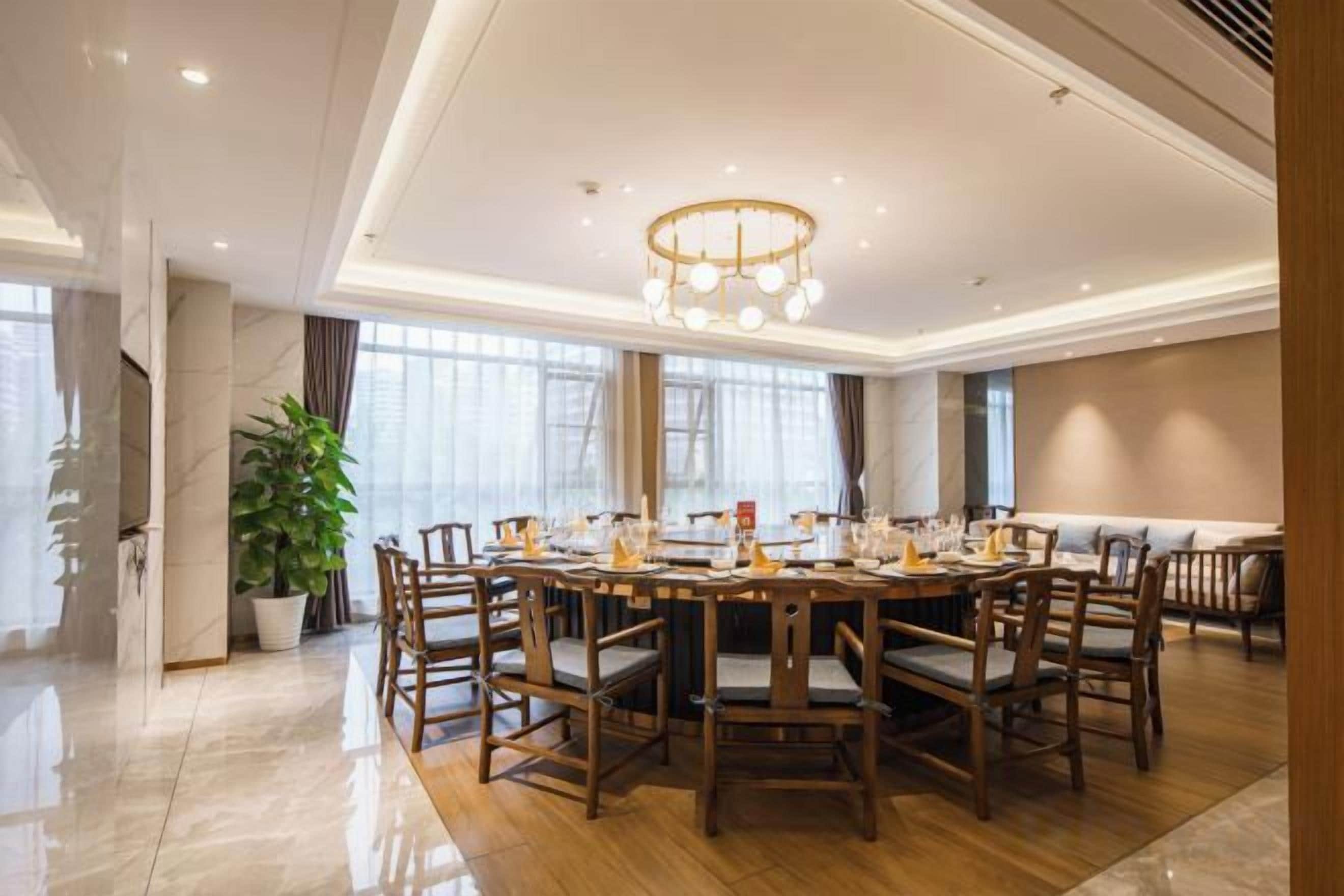 A spacious dining area with a round table and chairs, a chandelier, and large windows.