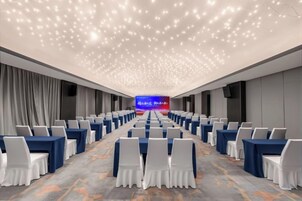 Un salón de conferencias con filas de mesas y sillas, una pantalla grande al frente y una alfombra estampada.
