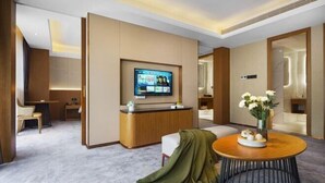 A modern hotel room with a flat-screen TV, a wooden reception desk, a round table with a vase of flowers, and a seating area with a chair and a bench. - Wellton City Hotel (Ganzhou Railway Station) (Ganzhou)