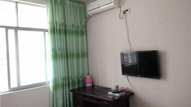 Une chambre dotée d’une fenêtre, d’un climatiseur, d’un téléviseur à écran plat fixé au mur et d’un bureau sur lequel se trouve une bouteille d’eau rose.