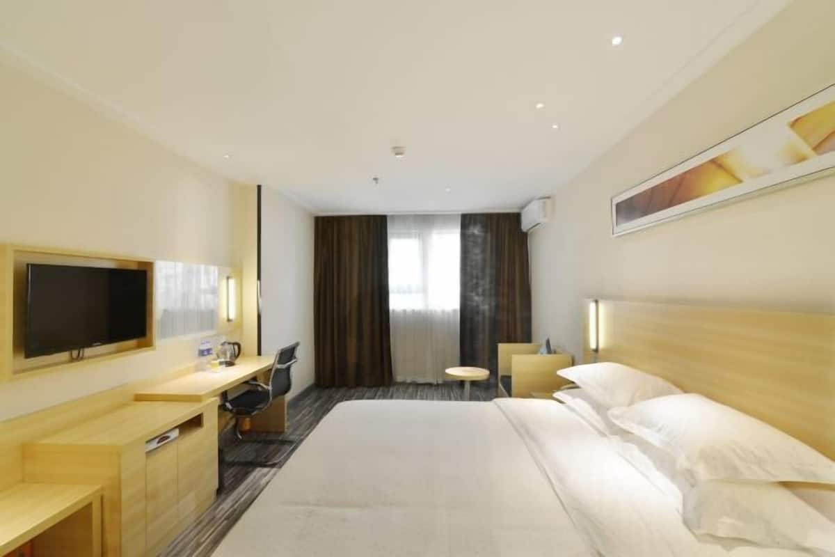 A hotel room with a bed, a desk, a chair, a TV, a small table, and a window with curtains.