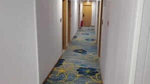 Een hotelgang met vloerbedekking met bloemenprint, witte muren en houten deuren.