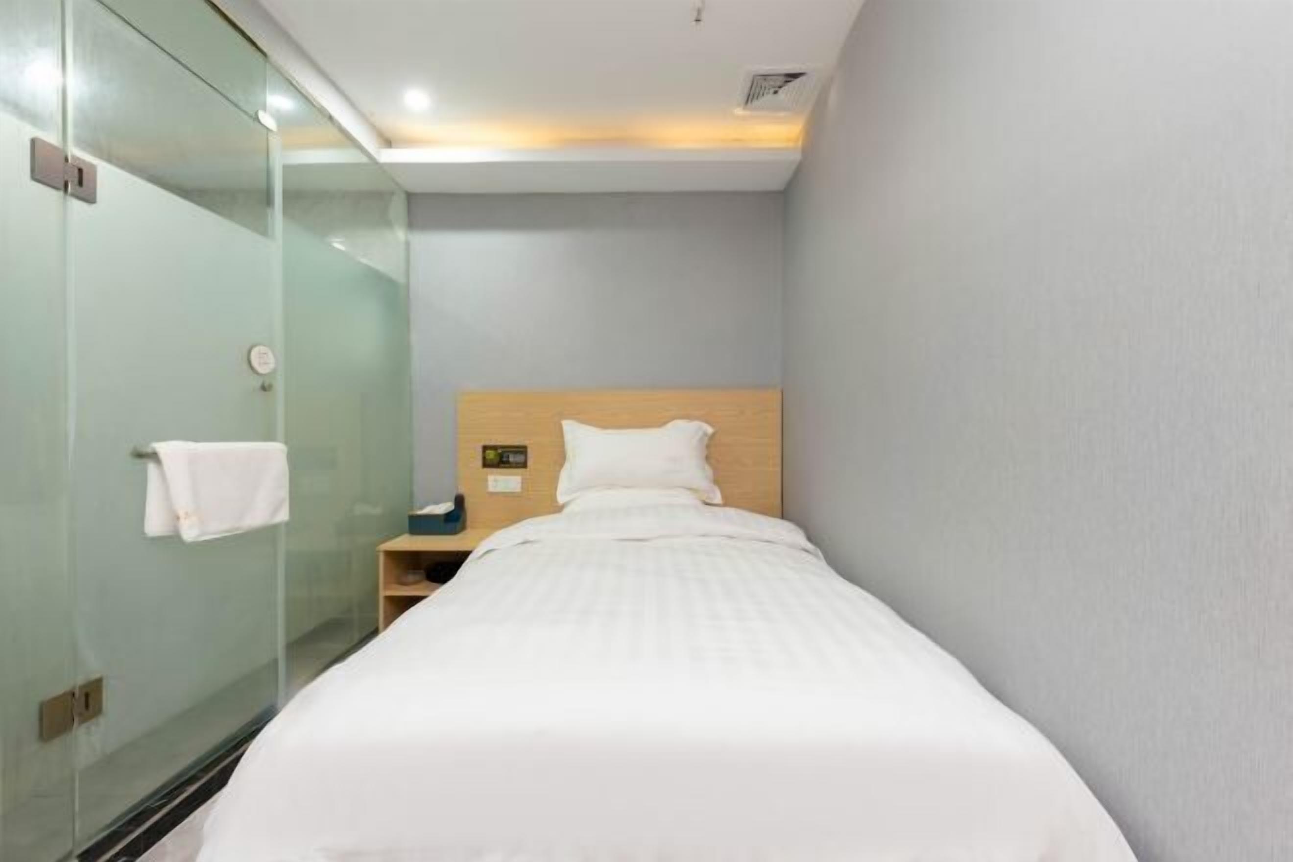 A narrow hotel room with a single bed, a bedside table, a nightstand, and a glass shower enclosure.
