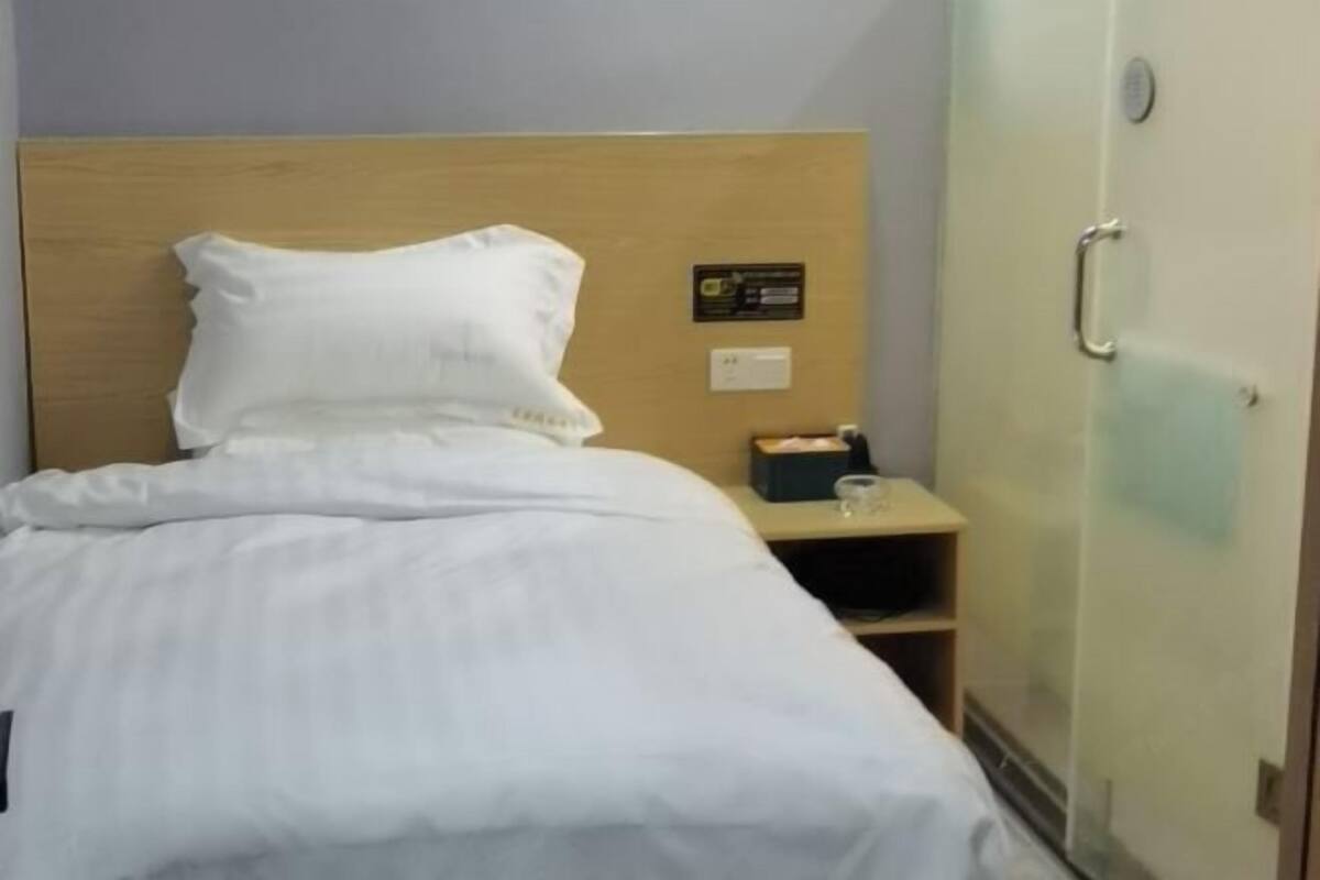 Een hotelkamer met een houten hoofdbord, een wit laken, een nachtkastje met een lamp en een klein plankje.
