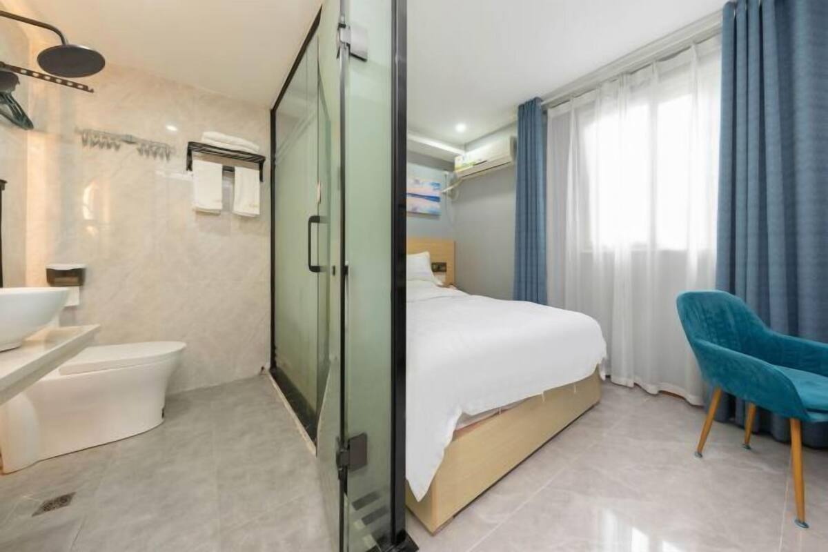 Een moderne hotelkamer met een glazen douchecabine, een wit wastafelmeubel, een toilet, een bed met een witte dekbedovertrek en een blauwe fauteuil.