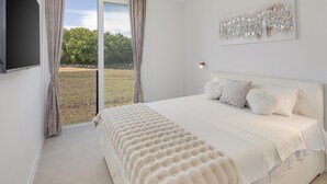 A bedroom with a large bed, a flat-screen TV, and a view of a grassy area through the window. - Luxurious home in Vrvari with swimming pool (Vrvari)
