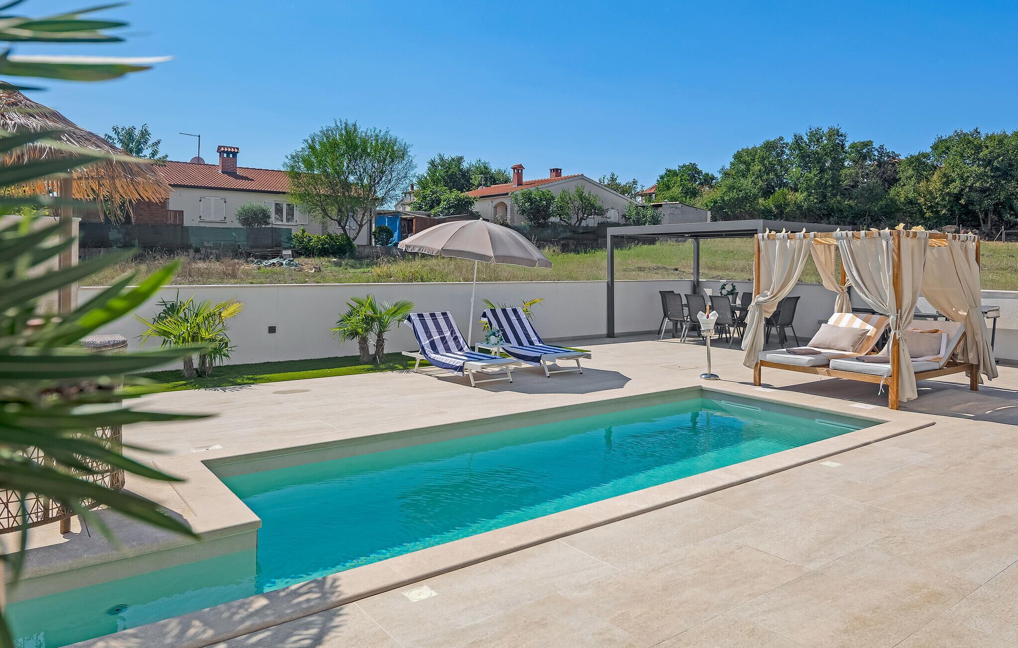 Une piscine dotée de chaises longues et d’un auvent, entourée d’un jardin et bordée de bâtiments en arrière-plan.
