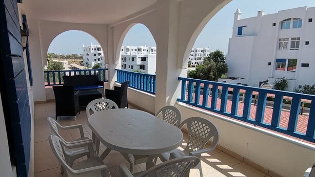 A balcony with a table and chairs, a grill, and a view of white buildings. - Family spacious apartment with sea view, AC, WiFi, 7 mins walking to the beach (M'diq)