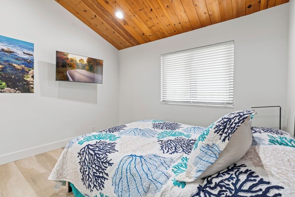 A bedroom with a bed, wooden ceiling, two wall-mounted paintings, and a window with blinds. - Gorgeous hillside home with ocean views! (Moss Beach)