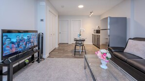 A living room with a TV, a sofa, a coffee table with flowers, a kitchen area, and a dining table. - 1 KING Modern Two Bedroom Secondary Suite in Airdrie Alberta (Airdrie)