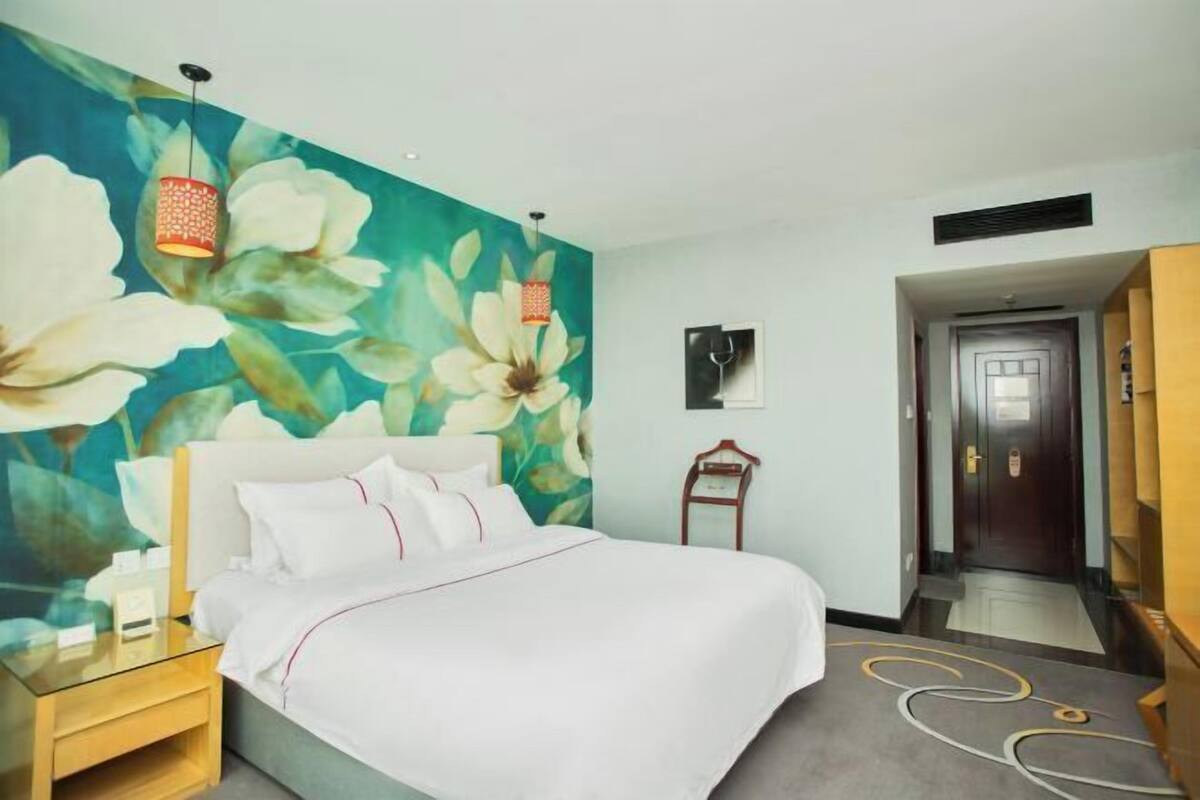 Een hotelkamer met een bed, een nachtkastje, een wand met een bloemmotief en een deur, en een gang.