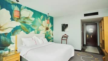 Hotellihuone, jossa on sänky, yöpöytä, kukka-aiheella koristeltu seinä, ovi ja käytävä.