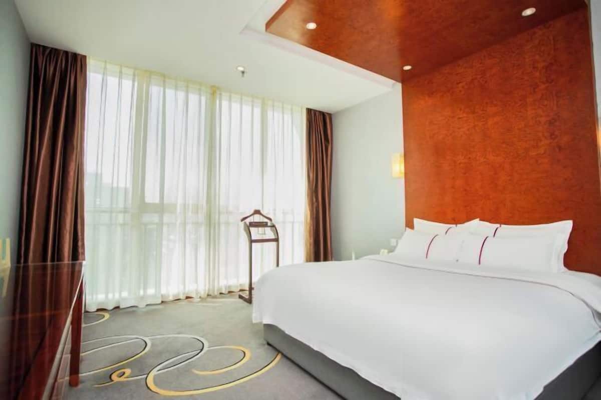 Een hotelkamer met een groot bed, een houten hoofdbord, een hangende kapstok en dunne gordijnen.