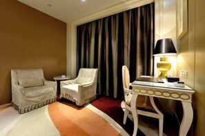 A hotel room with a large window, a desk with a lamp, two armchairs, and a patterned rug.