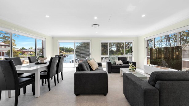A spacious living area with a dining table, black chairs, grey sofas, and large windows offering views of a residential area and a body of water.
