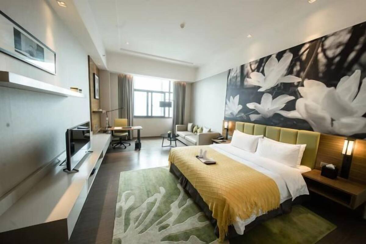 A hotel room with a large bed, a desk, a chair, and a sofa. There is a large window and a painting on the wall.