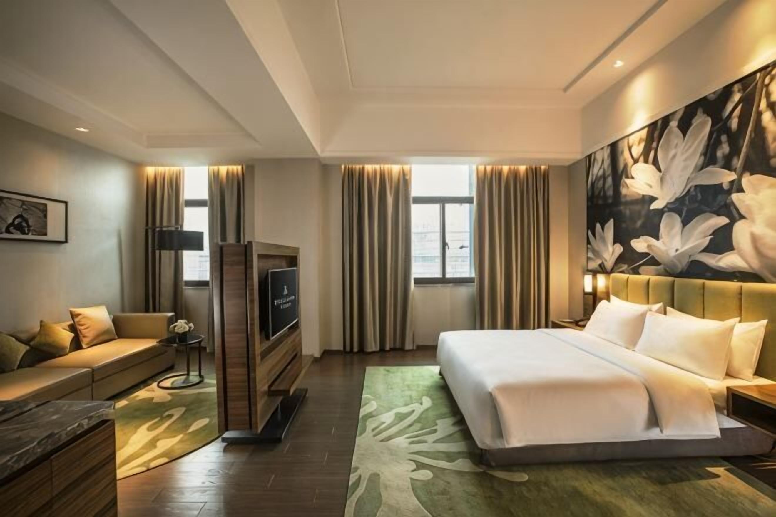A hotel room with a bed, a TV, a sofa, and a large floral artwork on the wall.