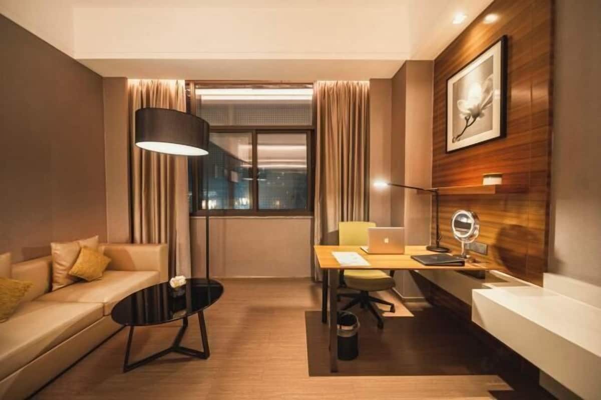 A modern hotel room with a sofa, a desk with a laptop, a lamp, a mirror, and a view of the city at night.