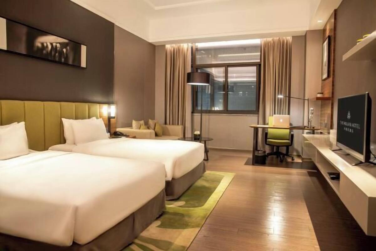 A hotel room with two beds, a desk, a chair, a TV, and a large window with curtains.