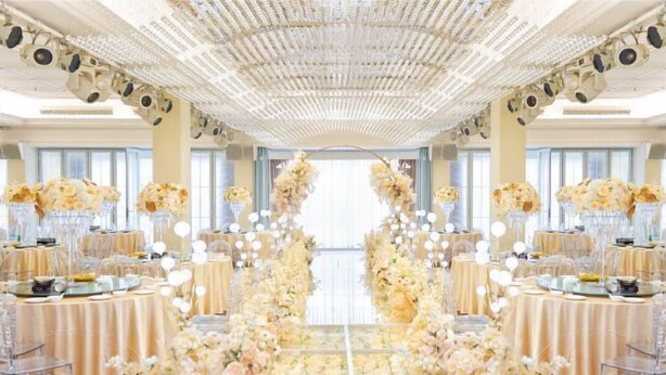 Un salón de banquetes con un pasillo central decorado con arreglos florales, mesas con sillas transparentes y un techo con diseño ornamental.