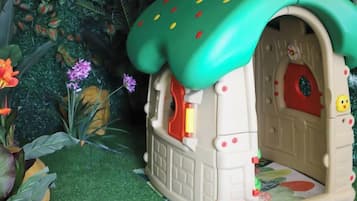 A small, enclosed playhouse with a green roof and a red door, surrounded by artificial plants.