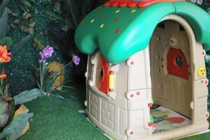 Una casita de juegos pequeña y cerrada, con techo verde y puerta roja, rodeada de plantas artificiales.
