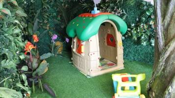 Una casita para animales con techo verde y puerta roja, rodeada de follaje artificial y un vehículo de juguete.