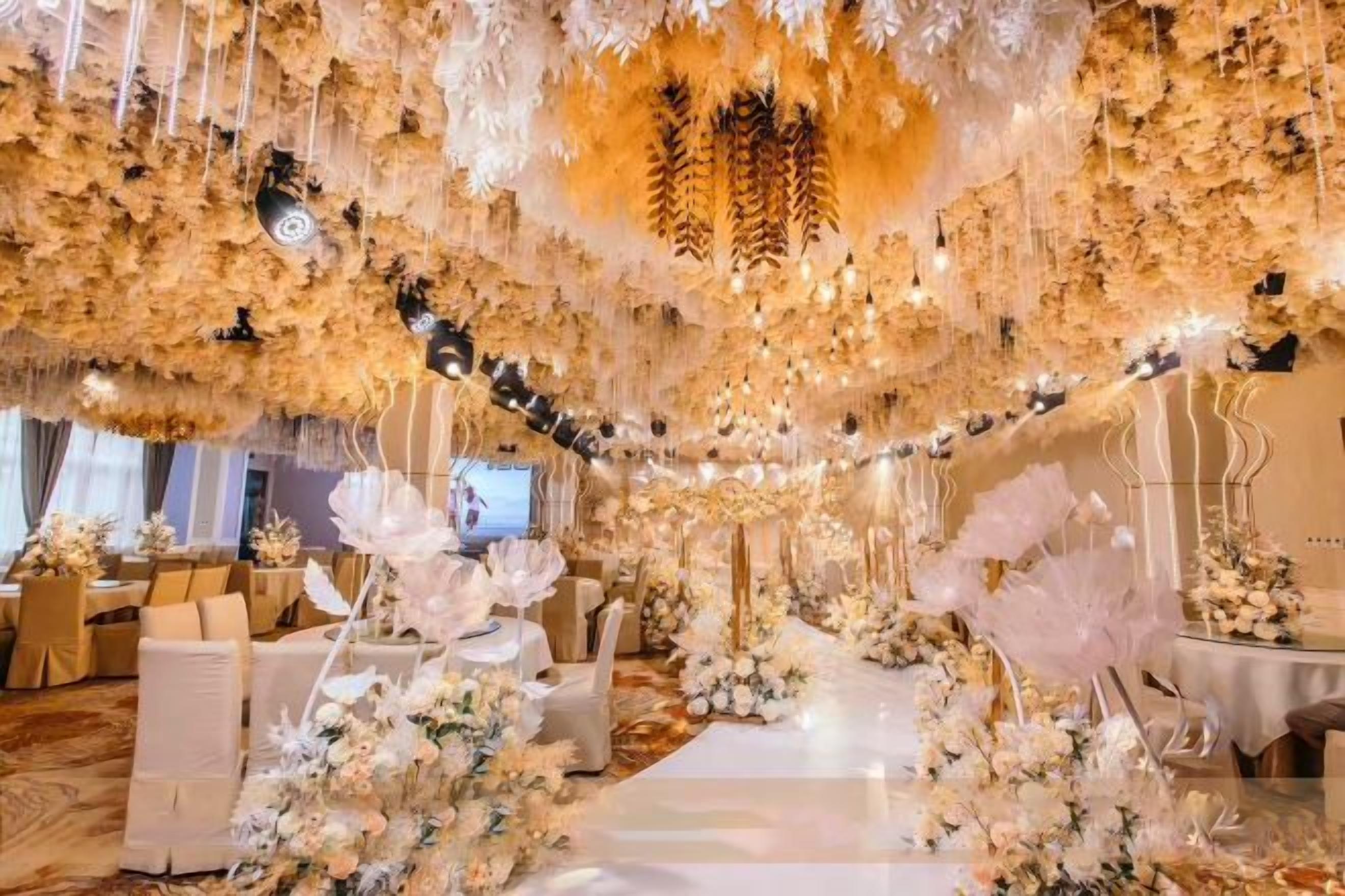 An indoor event space with a floral arrangement on the ceiling, tables with floral centerpieces, and a television screen in the background.